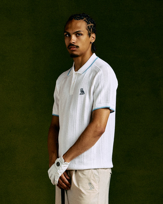 White open knit polo shirt with blue trim on collar and sleeves, worn by man against green background.