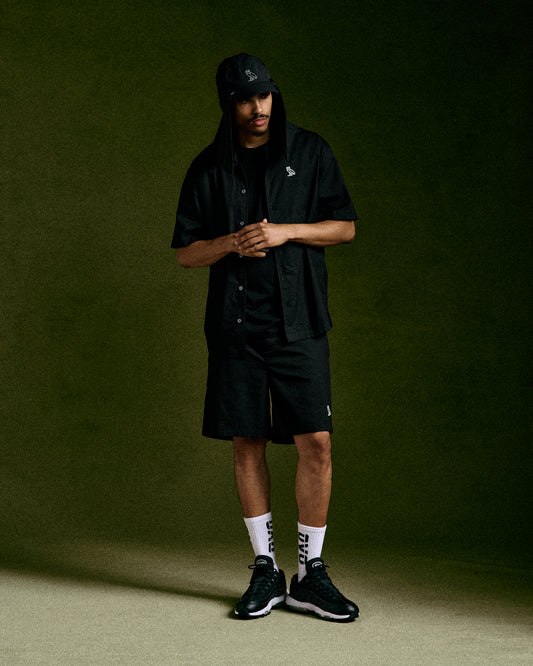 Black camp short with button-front closure and embroidered logo, displayed on model with matching cap and white socks.