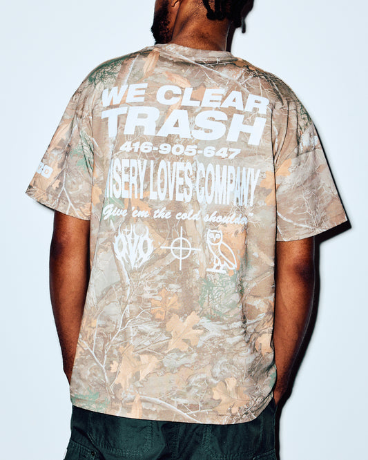Camouflage tee with white graphic text on back, shown on model from behind wearing dark pants.