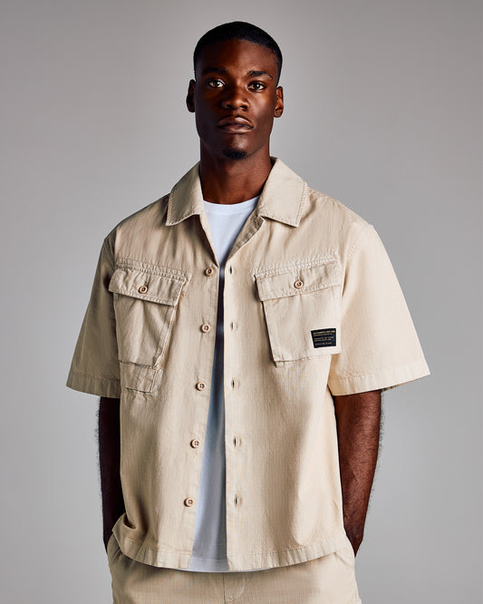 Cream-colored short-sleeve camp shirt with button front, chest flap pockets, and brand label on chest.