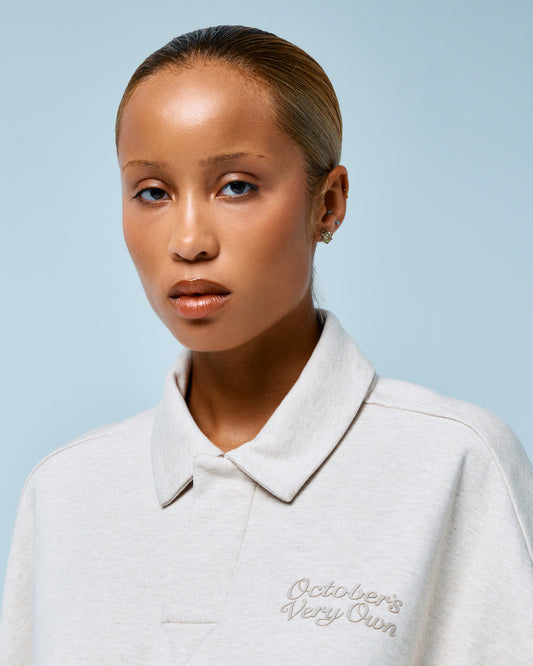 Heather oatmeal rugby polo with embroidered script text on chest and collared neckline.
