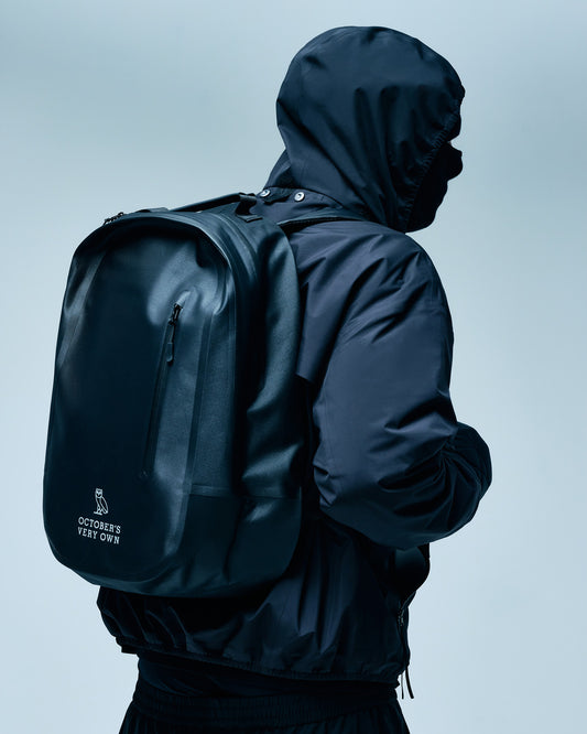 Black backpack with front zipper pocket and padded shoulder straps worn on a person's back.