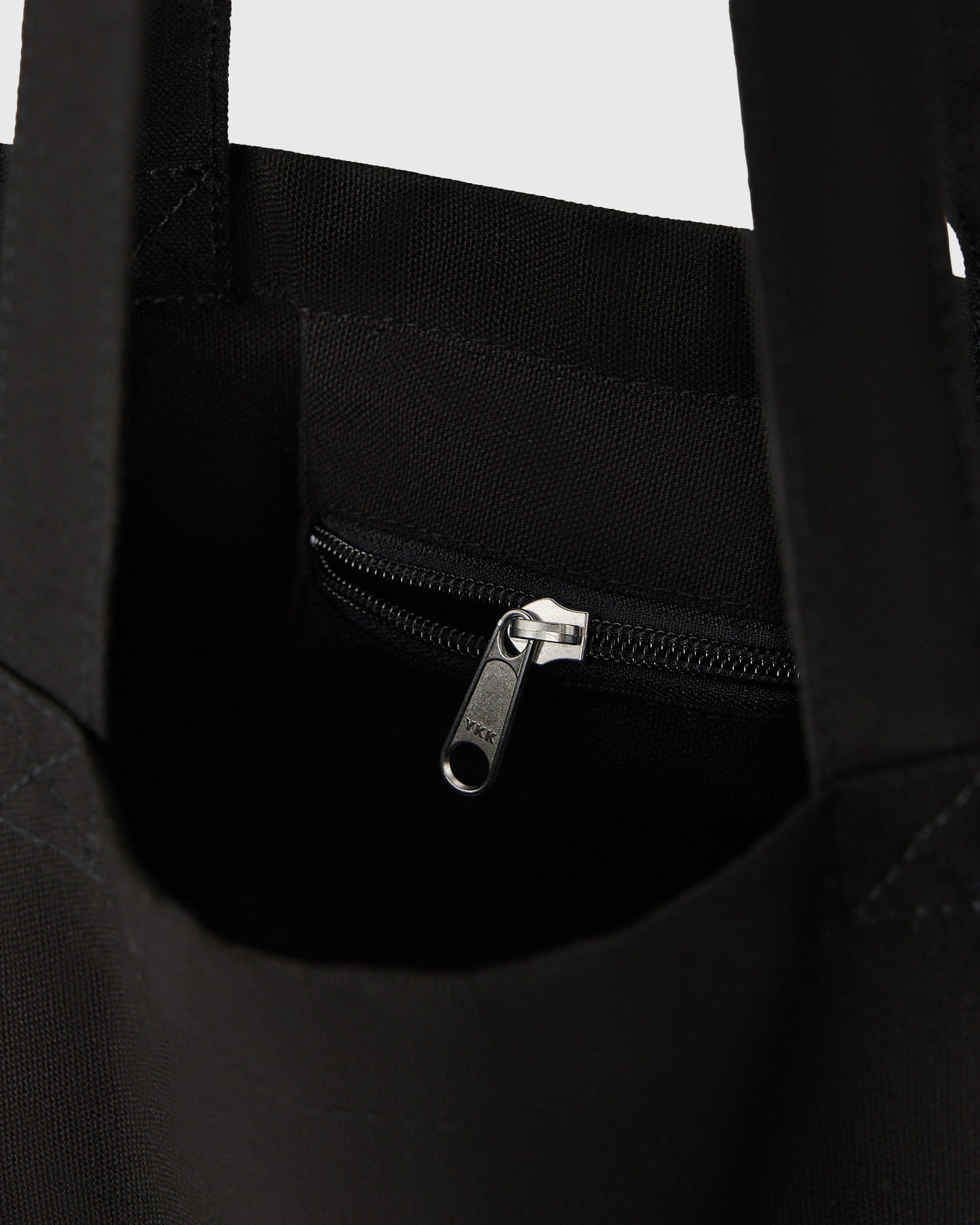 Black canvas tote bag with dual handles and silver metal zip closure on front pocket.