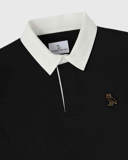 Black rugby polo with white collar and placket, gold owl embroidered on chest.