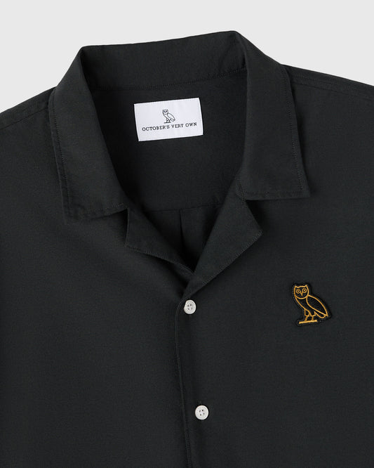 Black camp shirt with notched collar, white buttons, and gold owl embroidery on chest.