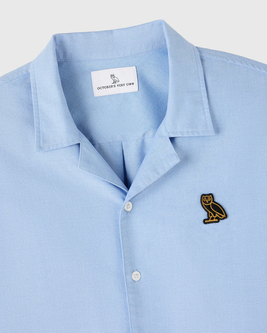 Light blue camp shirt with pointed collar, white buttons, and embroidered owl logo on chest.