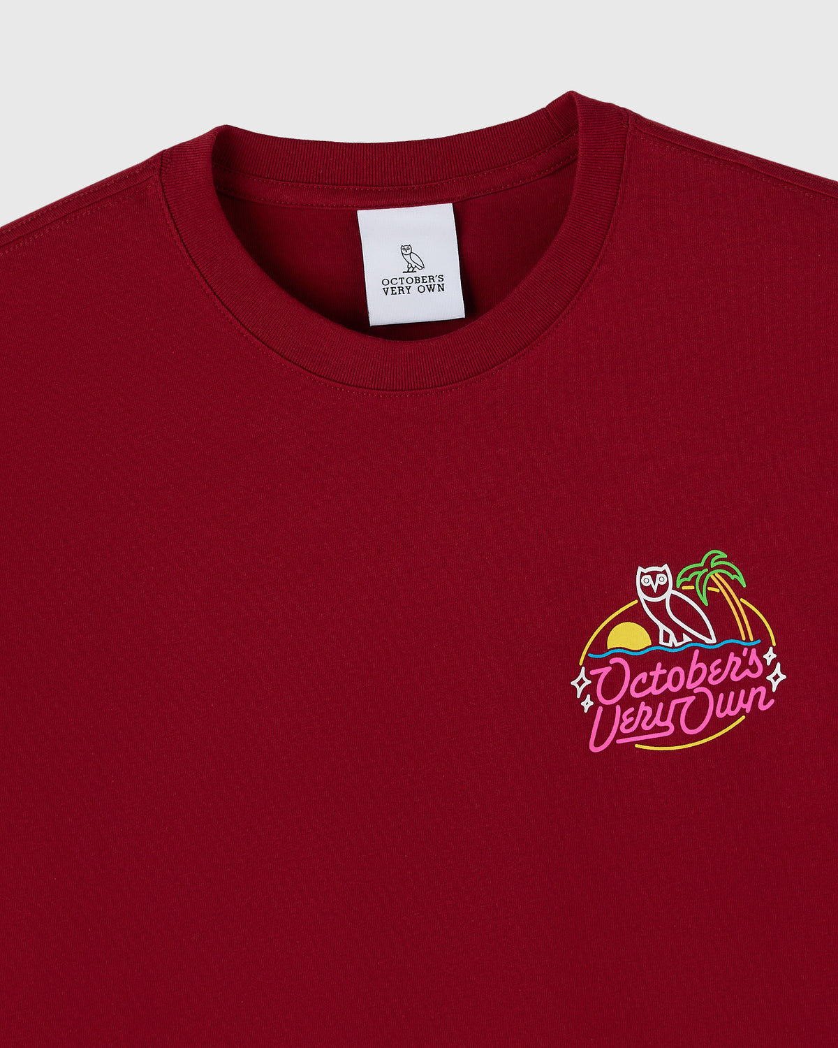 Deep red crewneck t-shirt with neon-style graphic featuring a figure, palm tree, and "October's Very Own" text on chest.