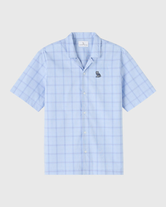 Light blue check camp shirt with short sleeves, white button front, and small circle wordmark emblem on chest.