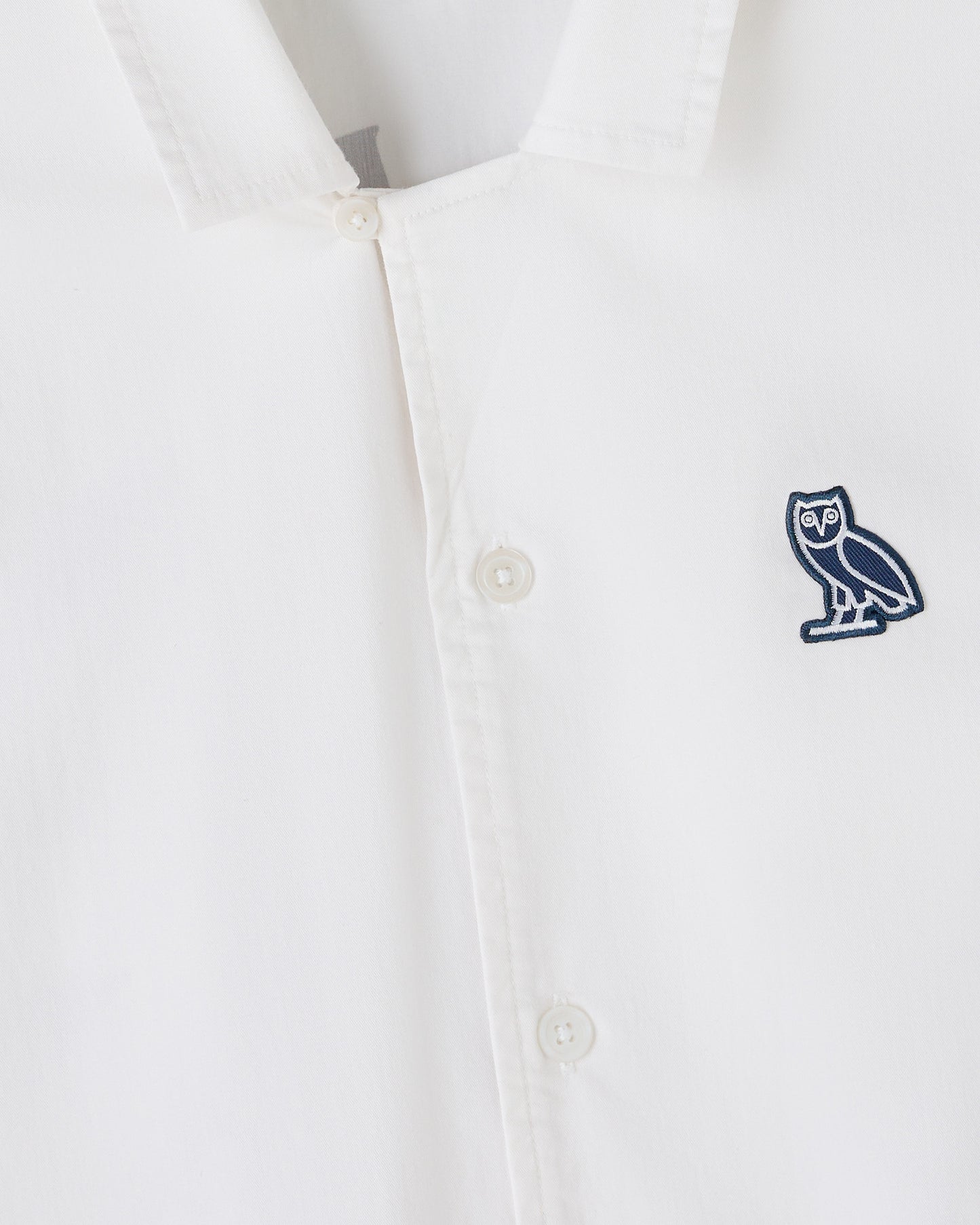 White camp shirt with button front closure and navy owl embroidered patch on chest.
