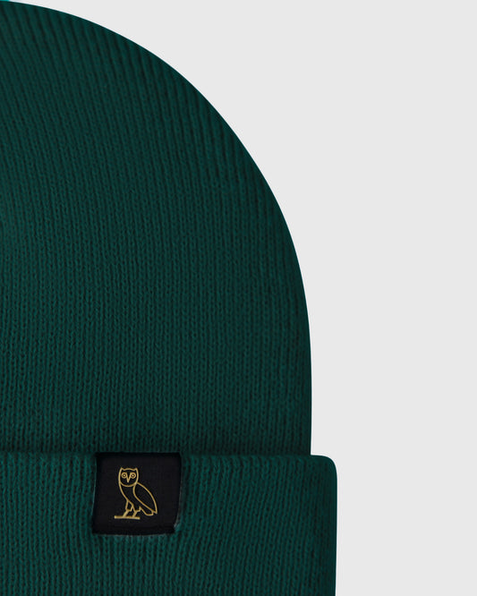 Midnight green ribbed knit beanie with black woven label featuring gold owl emblem.