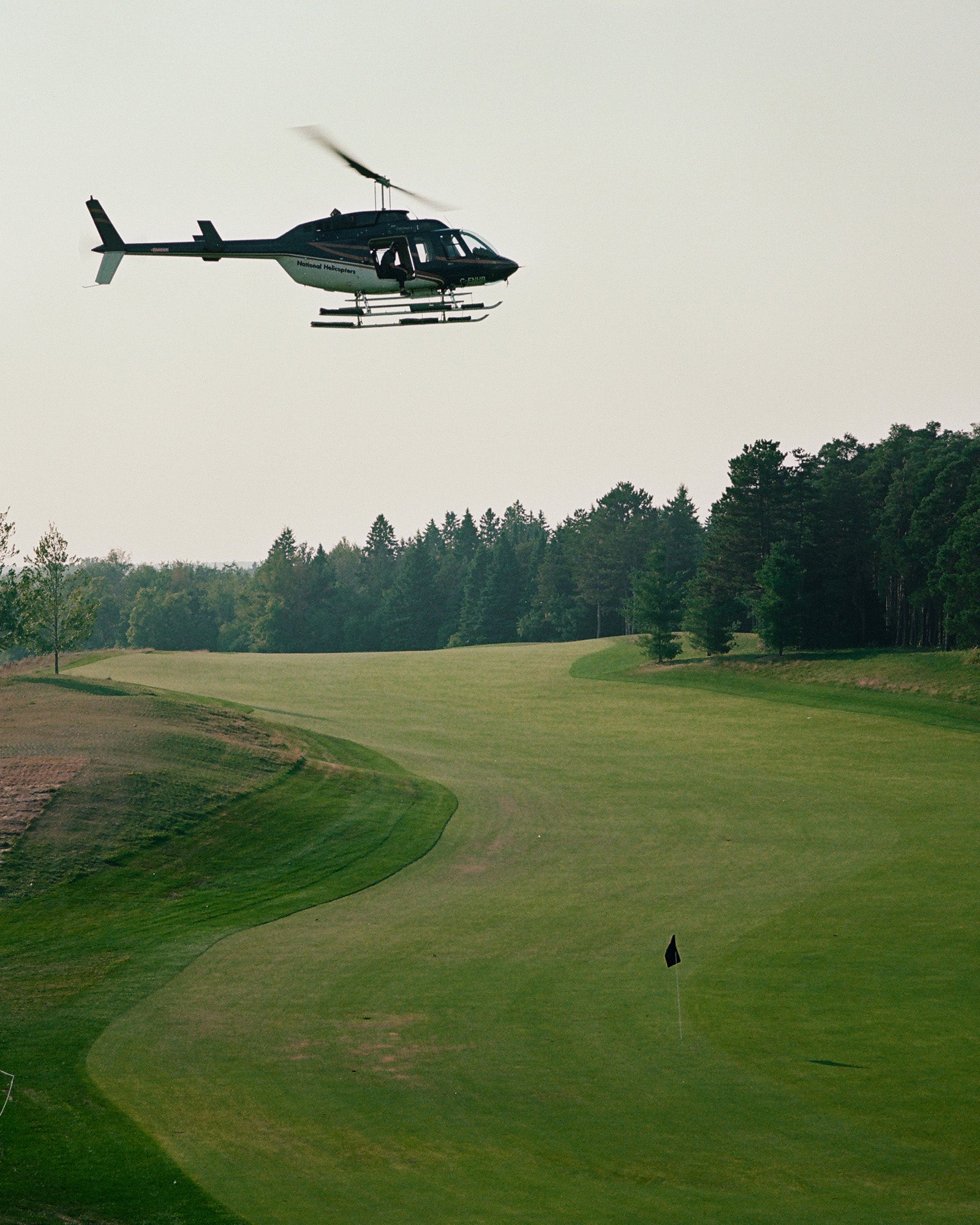 Helicopter flying over a golf course green with fairway, trees, and flag marker visible below.