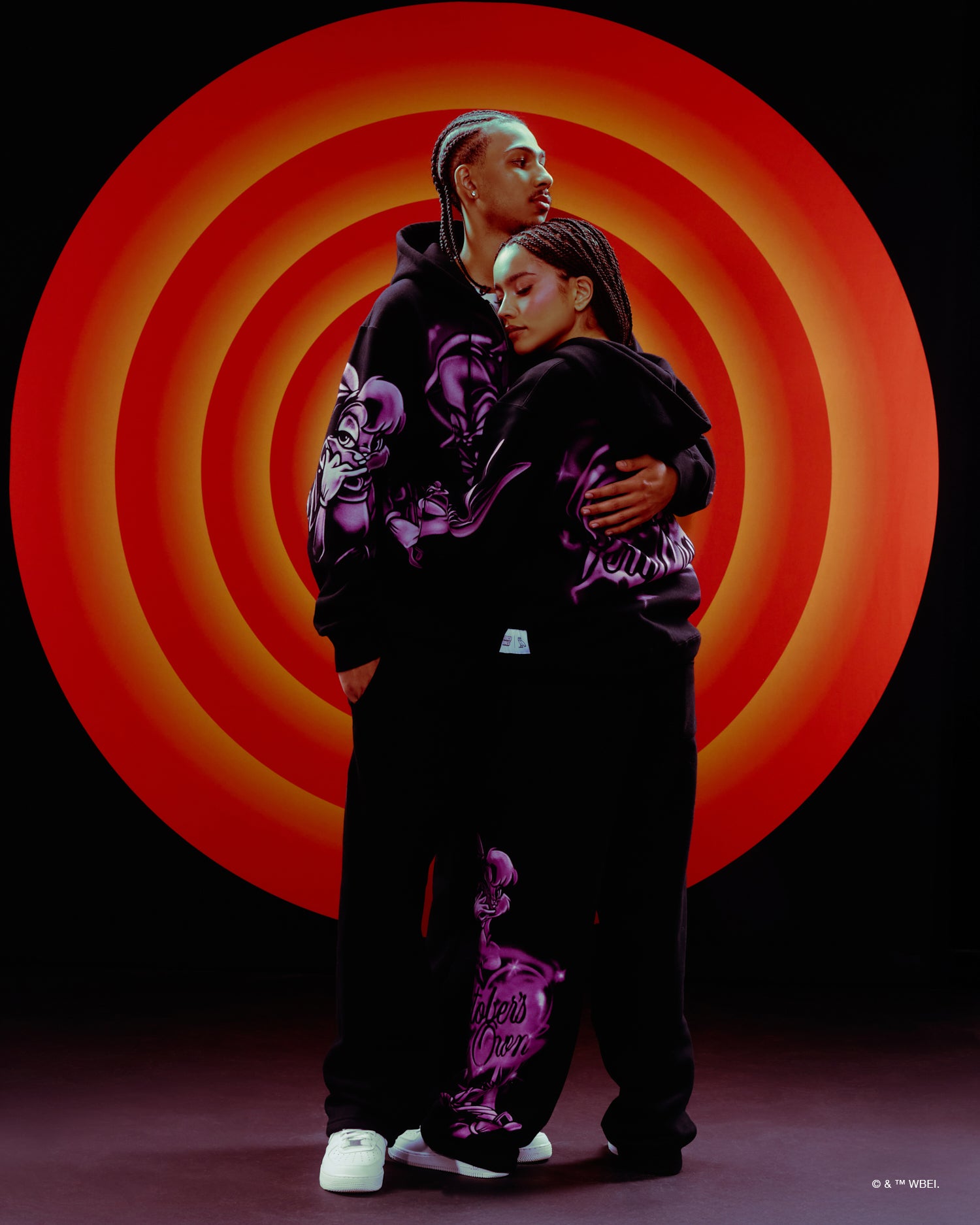 Two people wearing matching black October's Very Own x Looney Tunes and purple outfits with a red and orange circular background.
