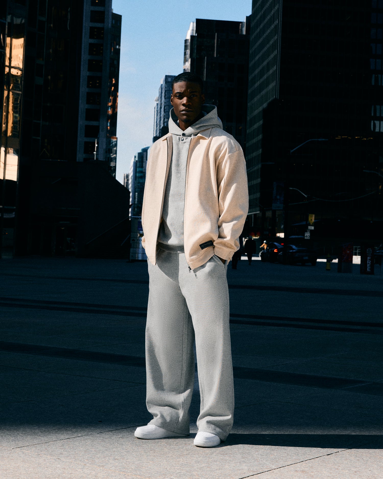 Person wearing a grey October's Very Own hoodie and light gray pants standing in an urban setting with tall buildings.