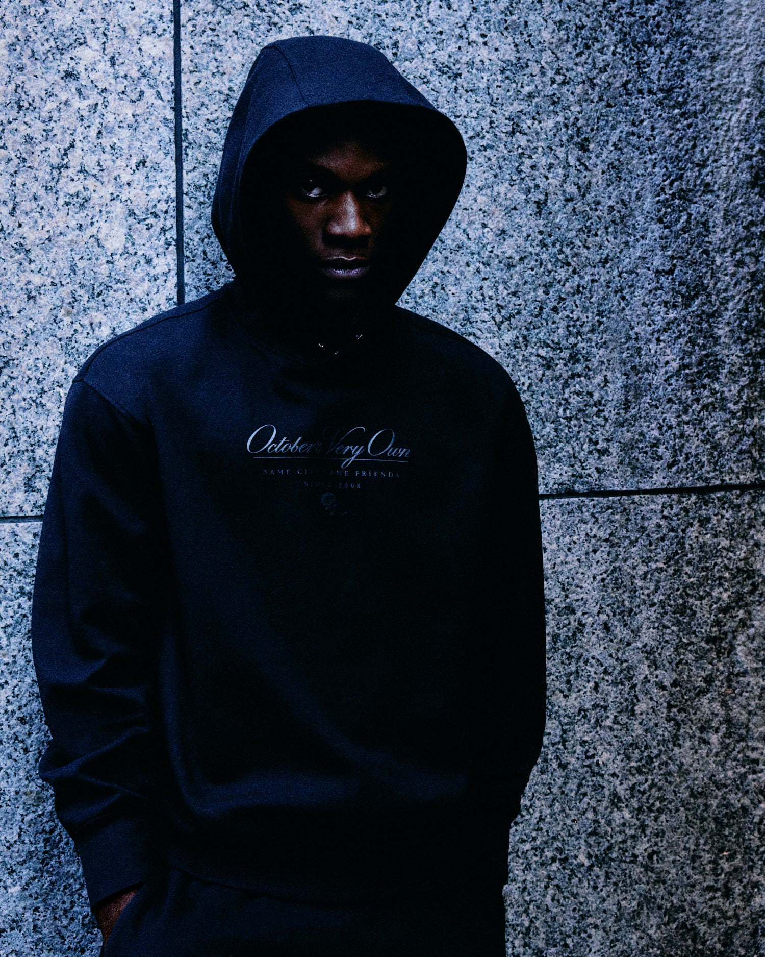 Person wearing a dark October's Very Own hoodie with text against a textured wall