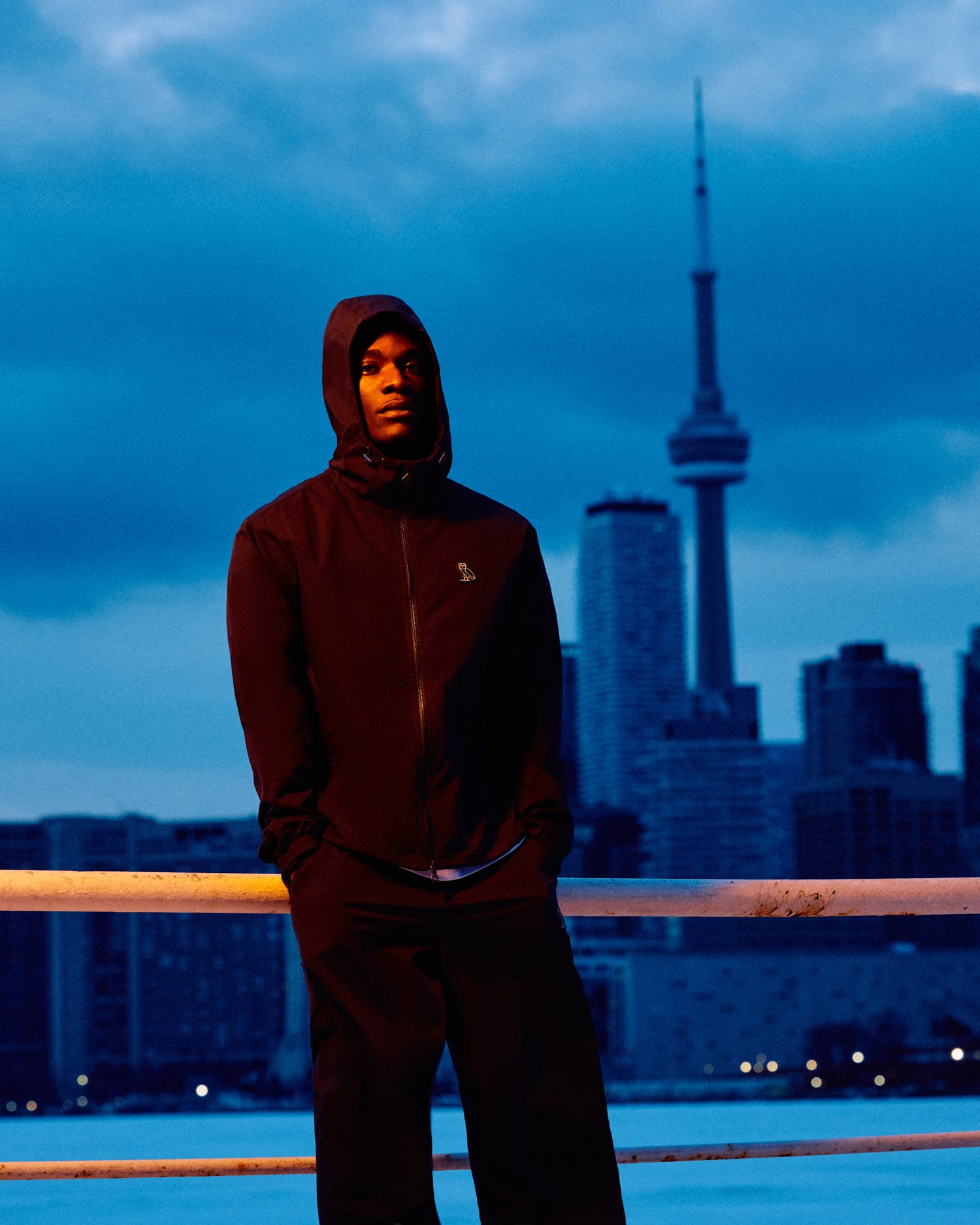 Person wearing a dark October's Very Own jacket with a hood in front of a city skyline at night.