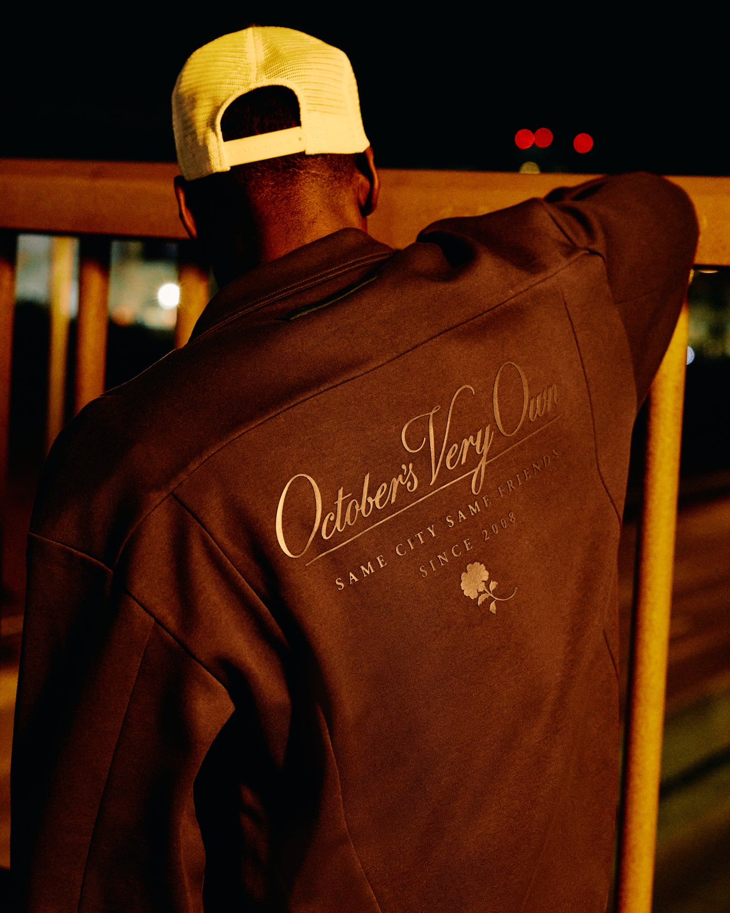Person wearing a brown jacket with 'October's Very Own' branding, standing on a wooden deck at night.