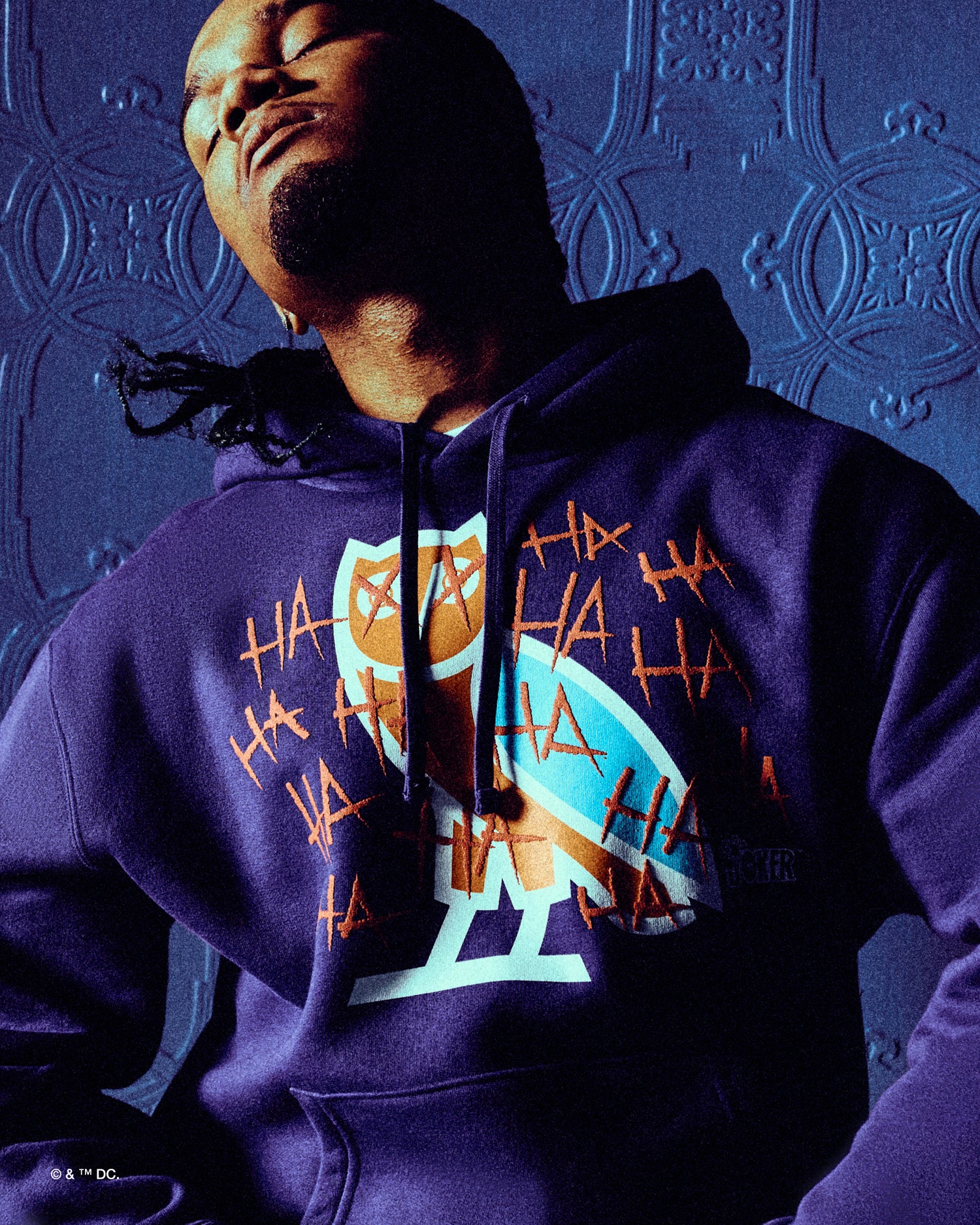 Person wearing a purple October's very Own x Batman x The Joker hoodie with a colorful logo against a textured blue background
