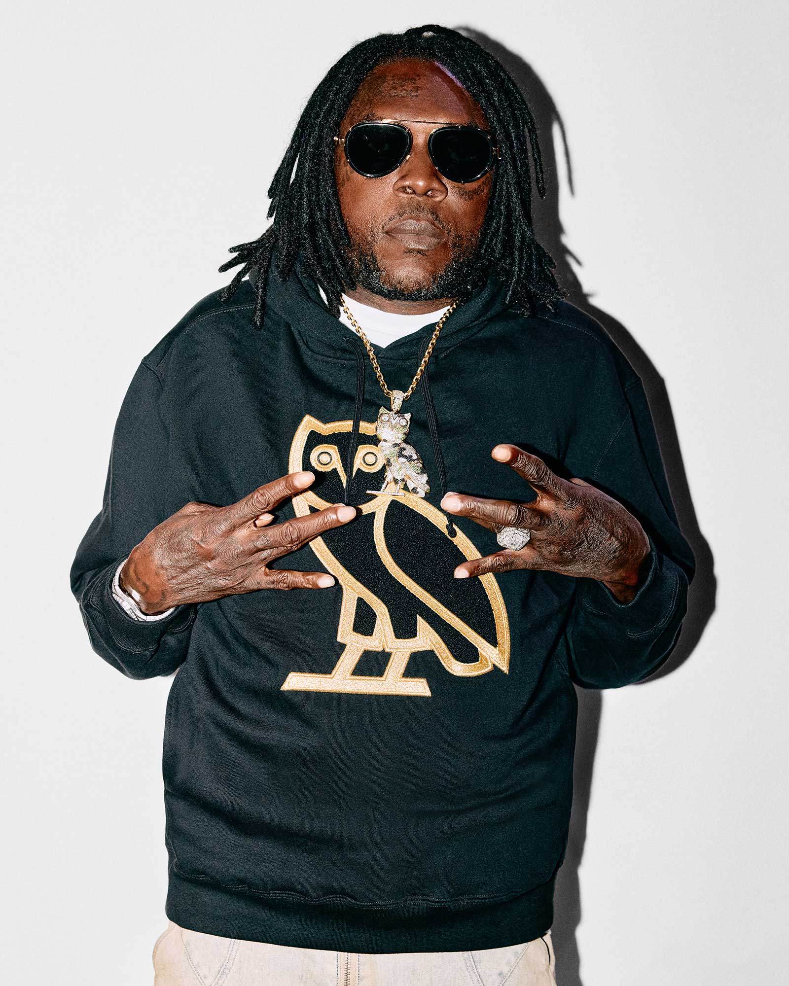 Vybz Kartel wearing a October's Very Own black hoodie with a gold logo, sunglasses, and jewelry on a white background