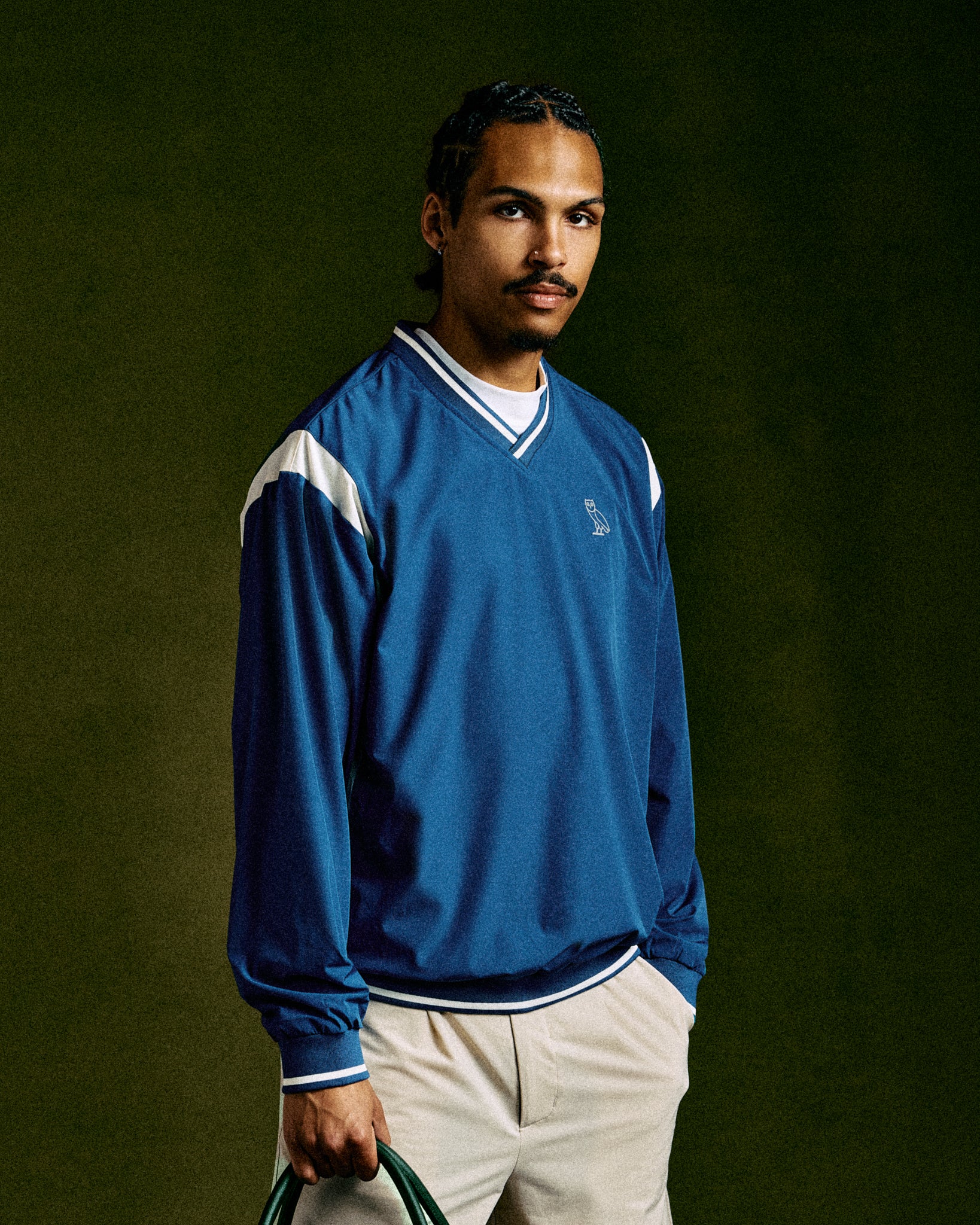 Deep denim V-neck pullover with white and navy stripe trim, worn with cream pants against green background.