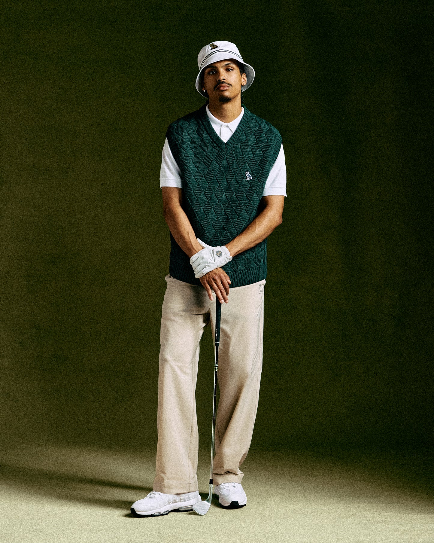 Midnight green basketweave sweater vest with V-neck worn over white t-shirt, paired with cream pants and golf accessories.
