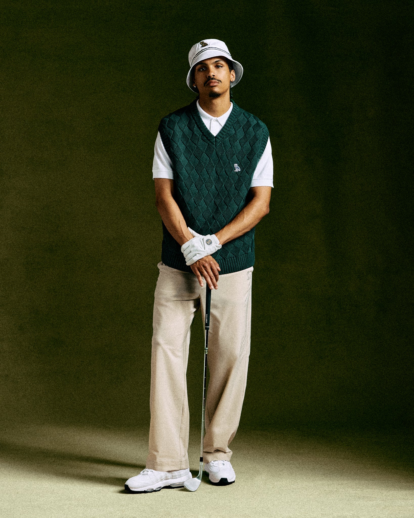 Midnight green basketweave sweater vest with V-neck worn over white t-shirt, paired with cream pants and golf accessories.