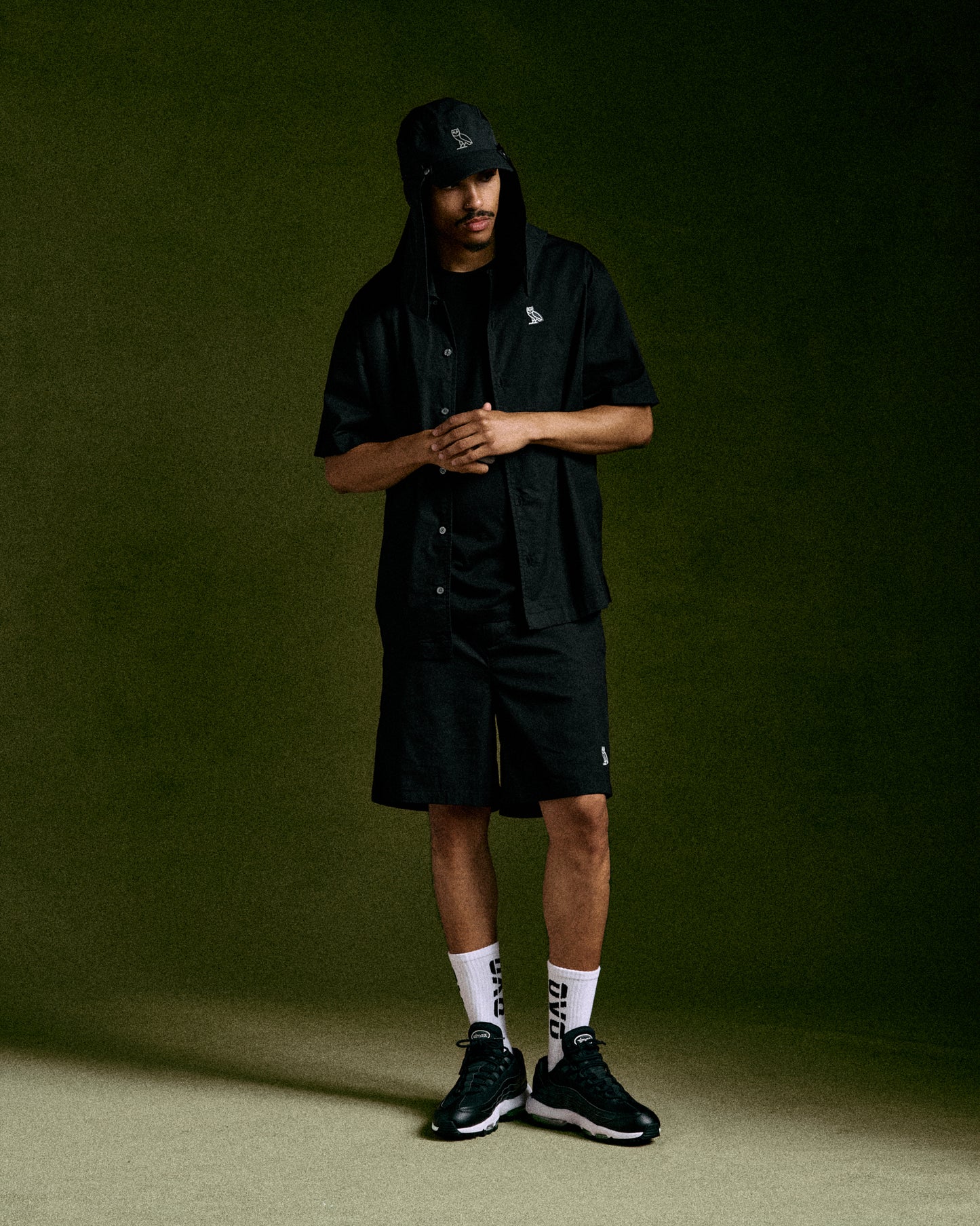 Black camp short with button-front closure and embroidered logo, displayed on model with matching cap and white socks.