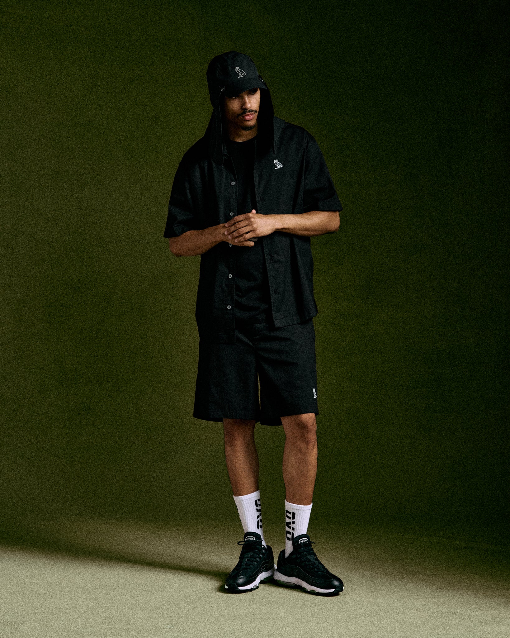 Black camp short with button-front closure and embroidered logo, displayed on model with matching cap and white socks.