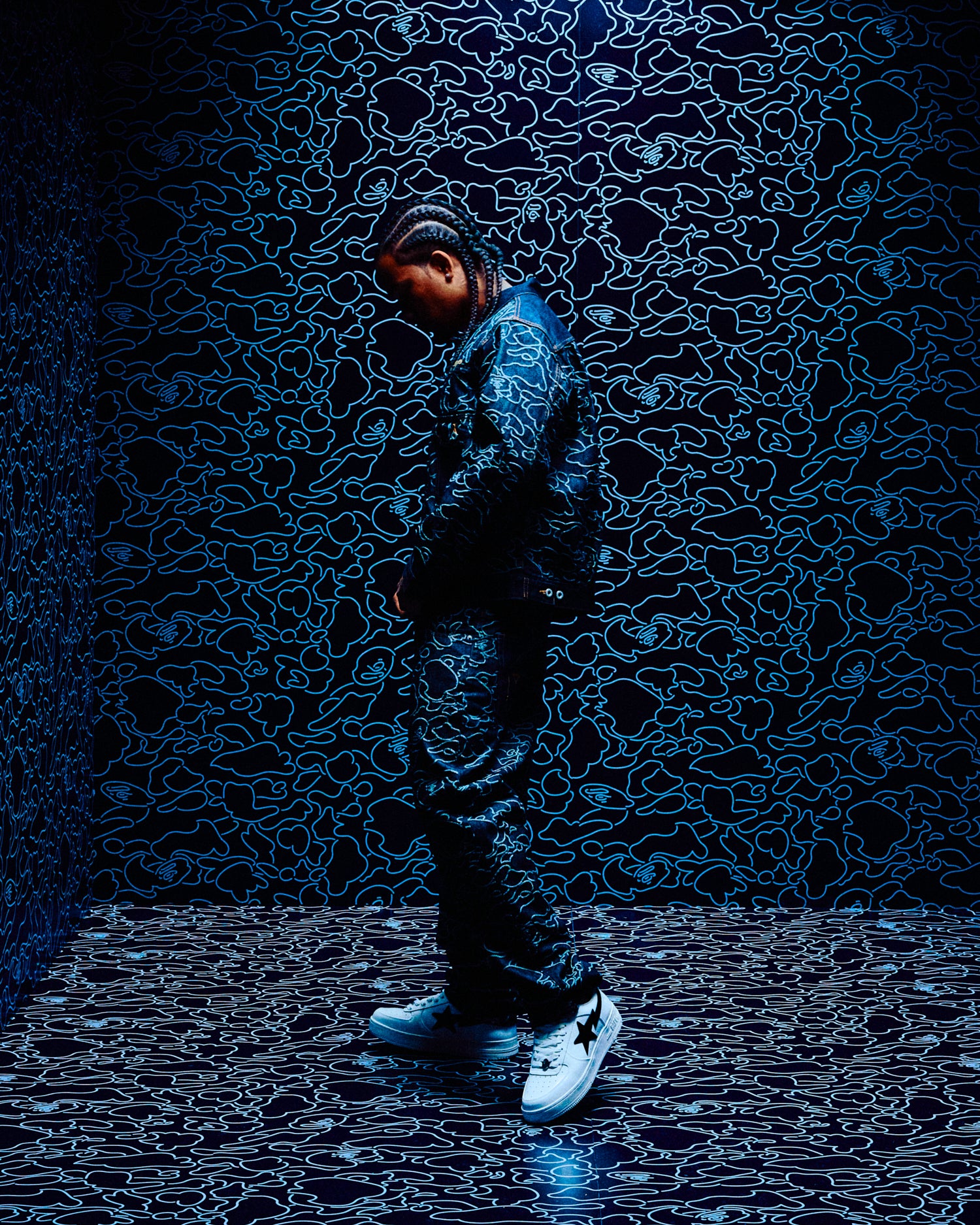 Person standing on a reflective surface with a dark, patterned background in October's Very Own x Bape Clothing
