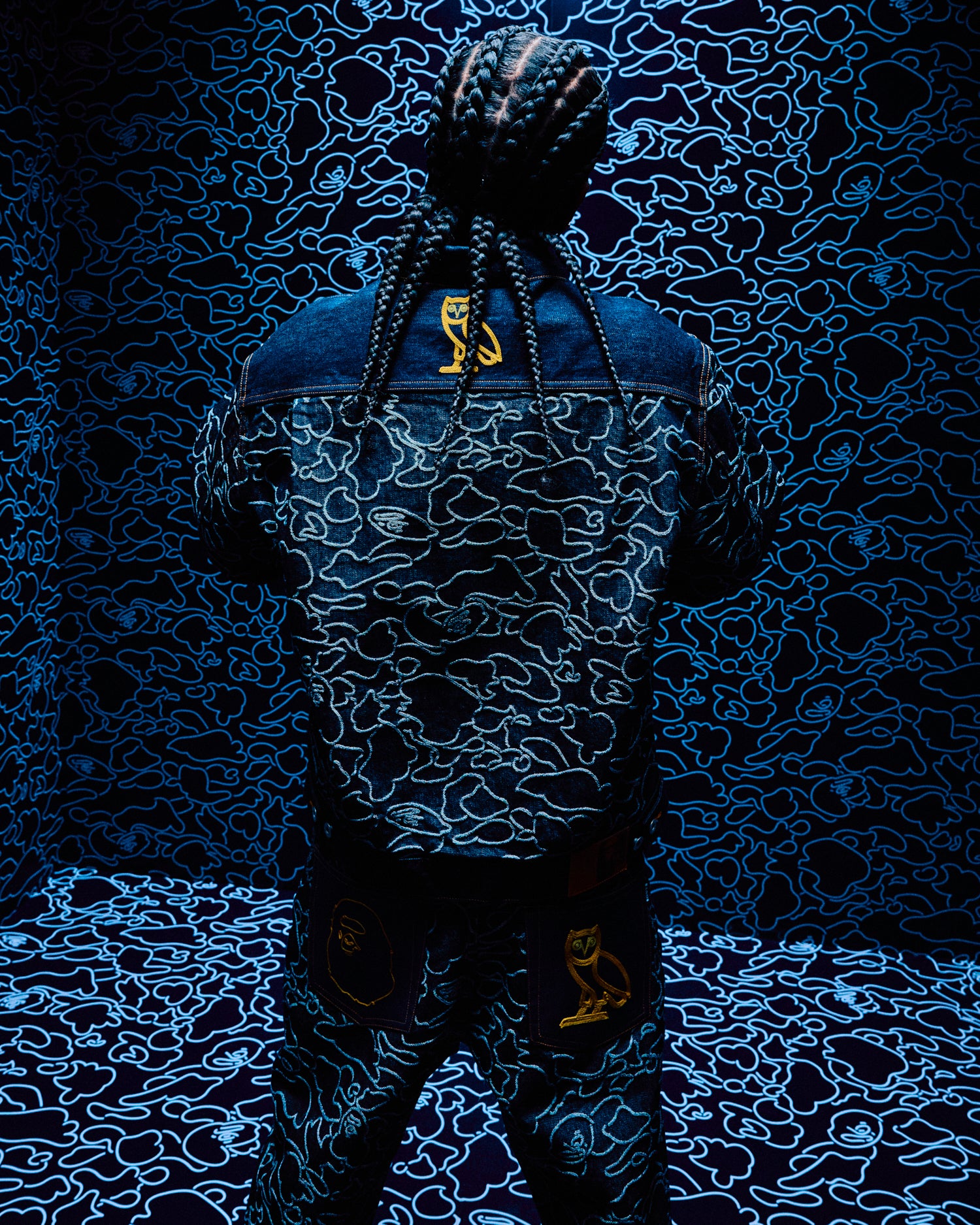 Person wearing a denim jacket with a logo against a October's Very Own x Bape dark, patterned background