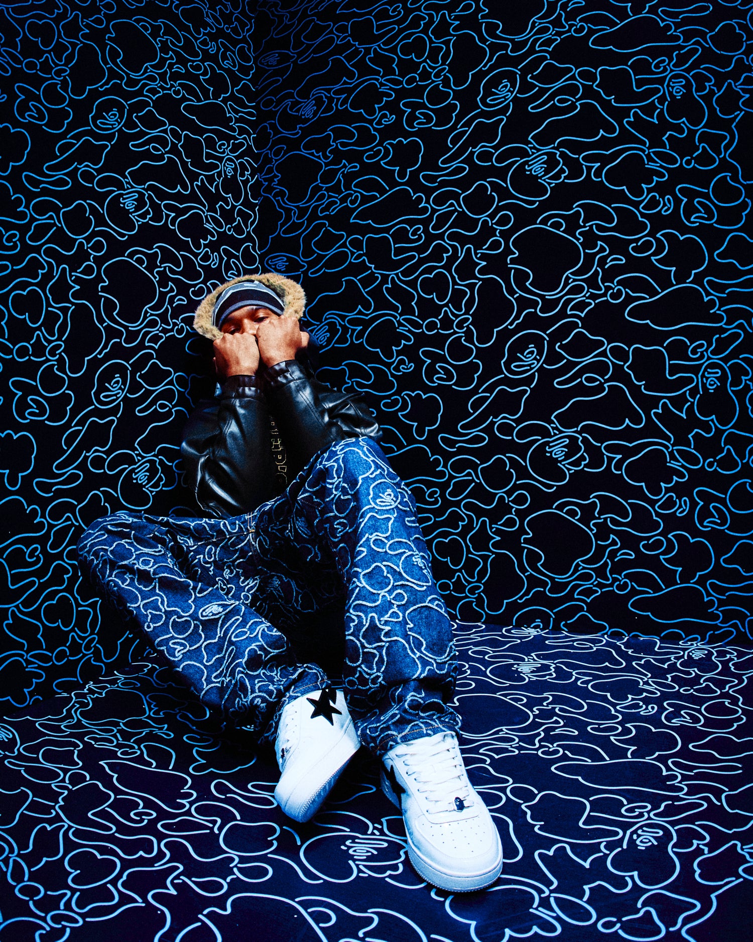 Person sitting on a dark patterned floor wearing October's Very Own x Bape black jacket, blue pants, and white shoes.