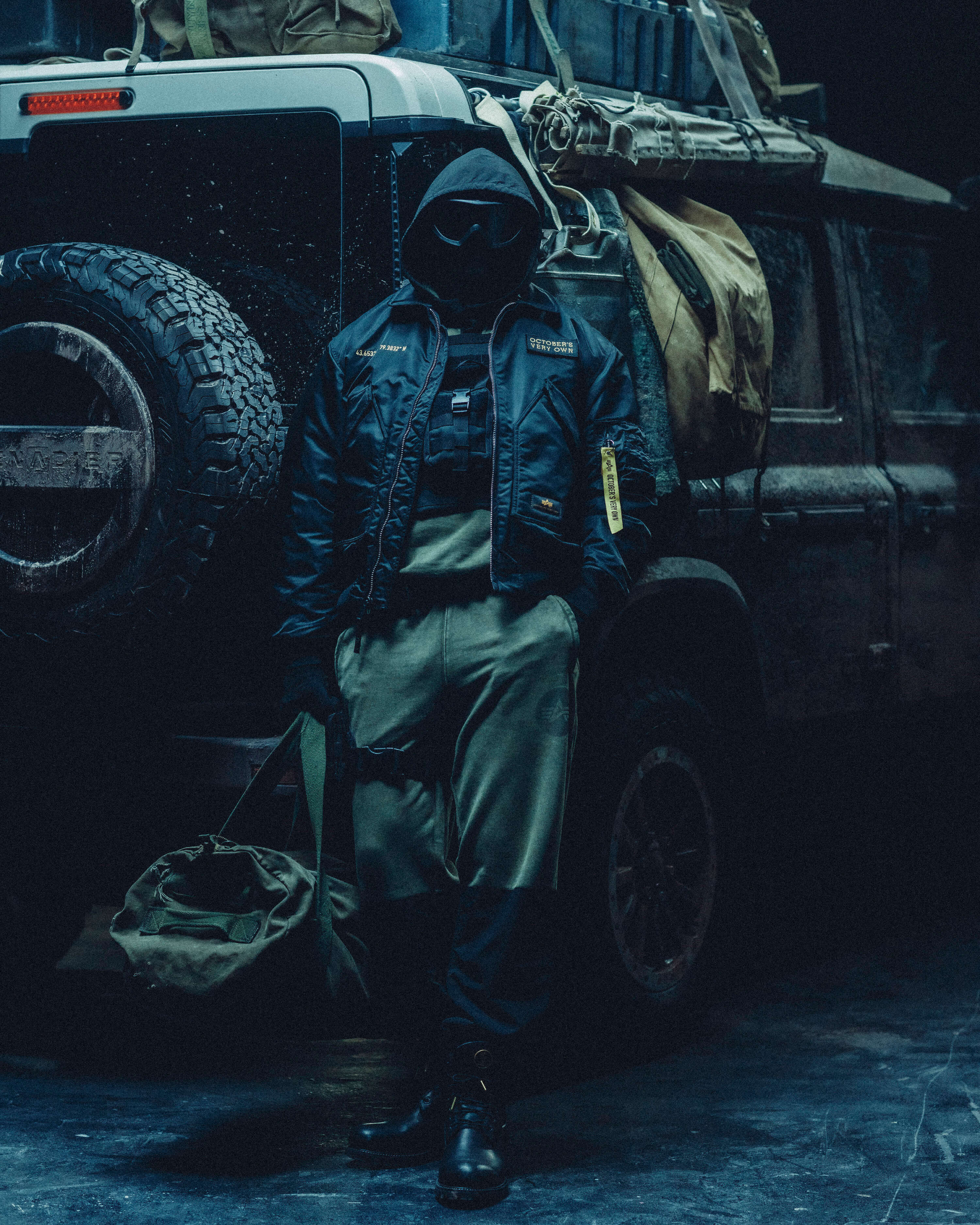 Person in October's Very Own x Alpha Industries gear standing next to a military vehicle in a dark setting
