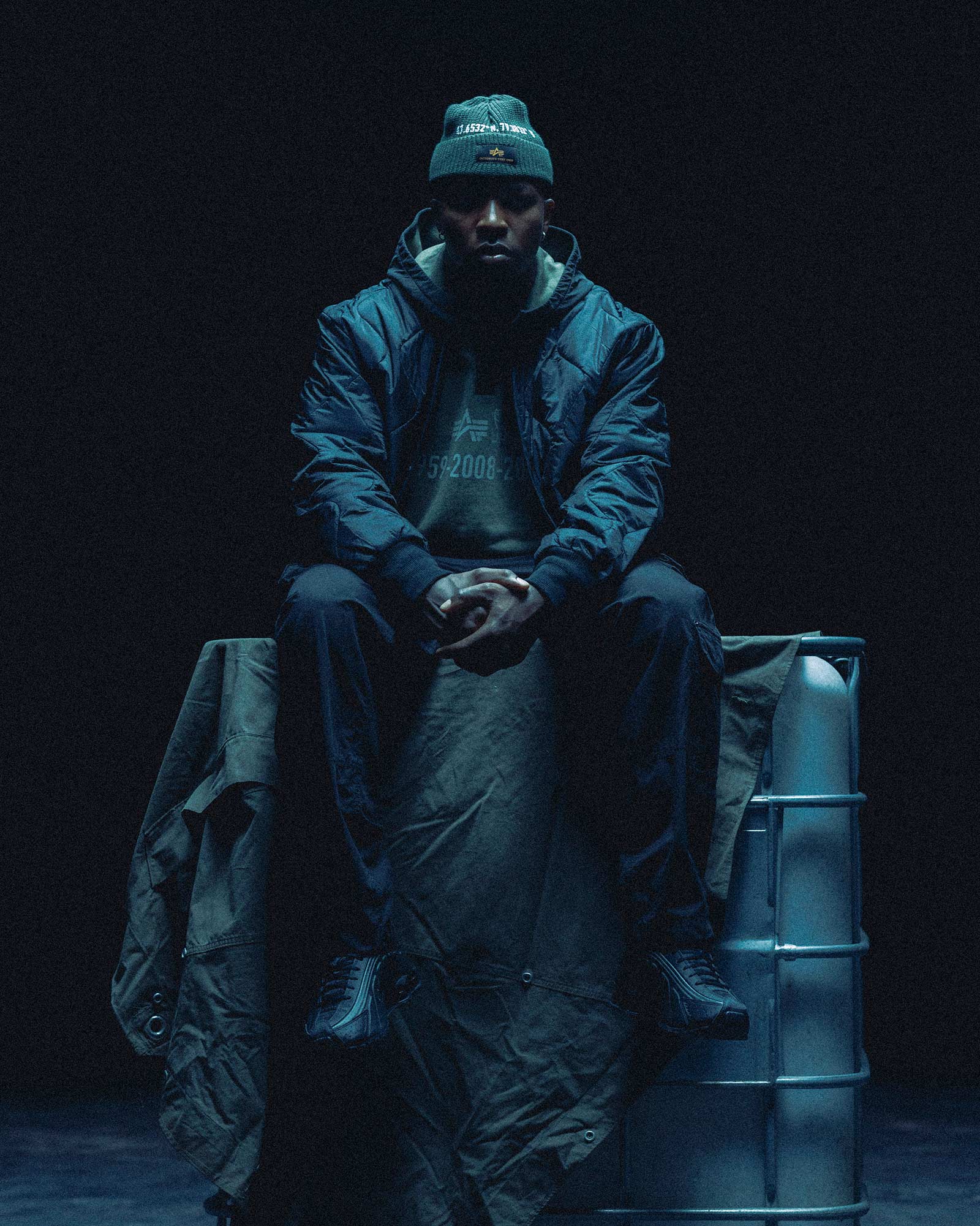 Person wearing a October's Very Own x Alpha Industries outfit sitting on a metal crate against a black background