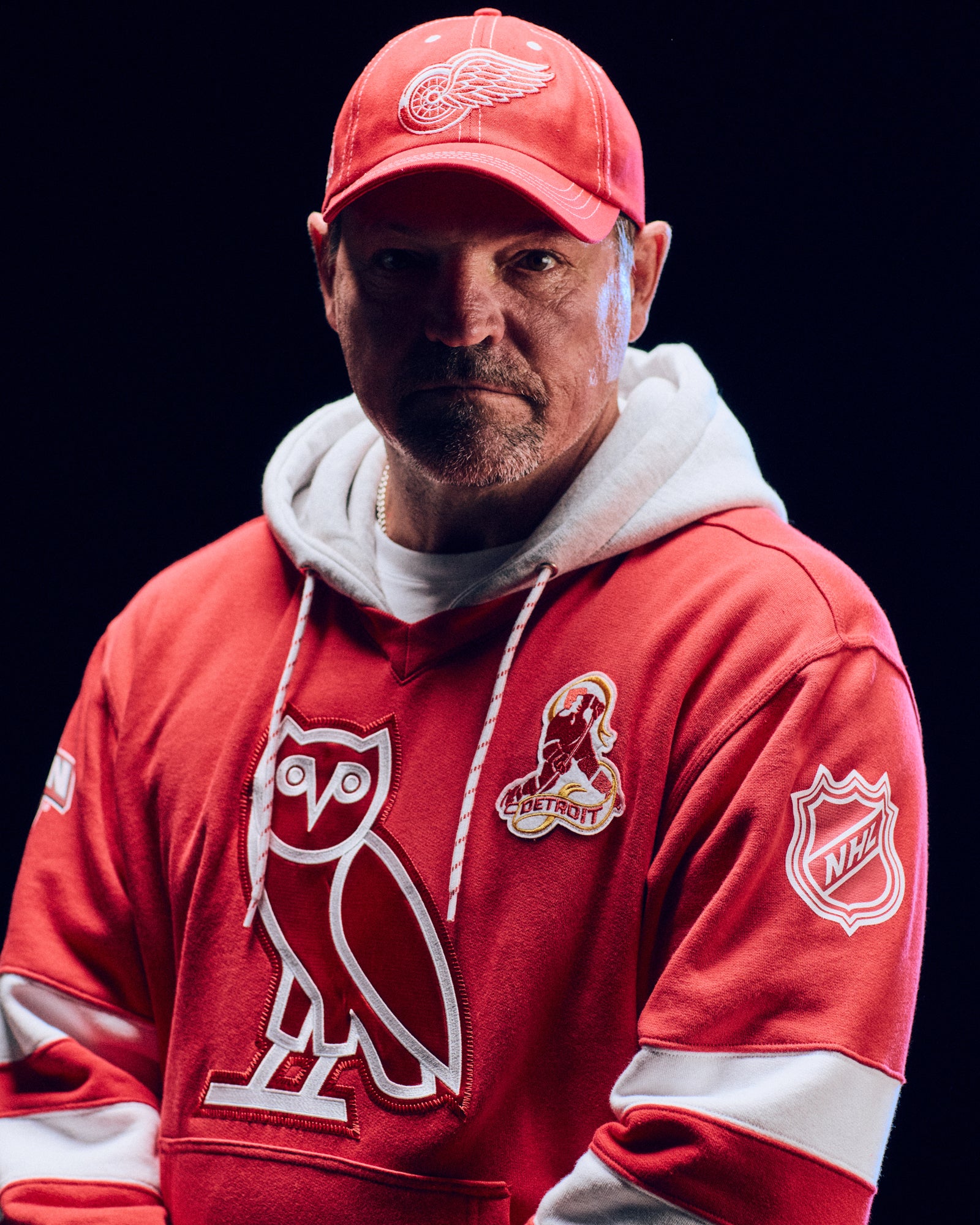 Person wearing a red October's Very Own x NHL  hoodie with logos and a red cap with a logo on a black background