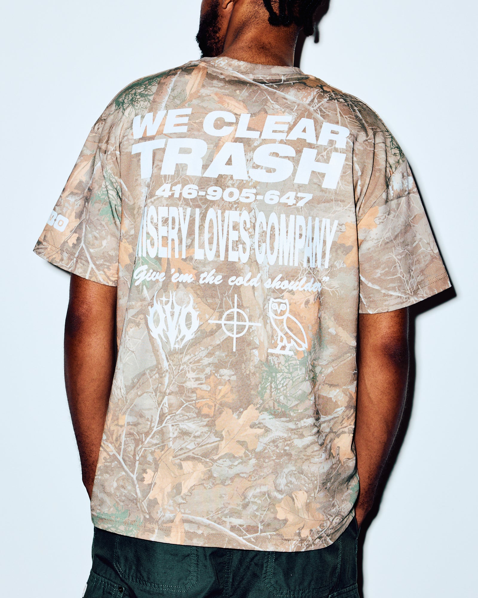 Camouflage tee with white graphic text on back, shown on model from behind wearing dark pants.