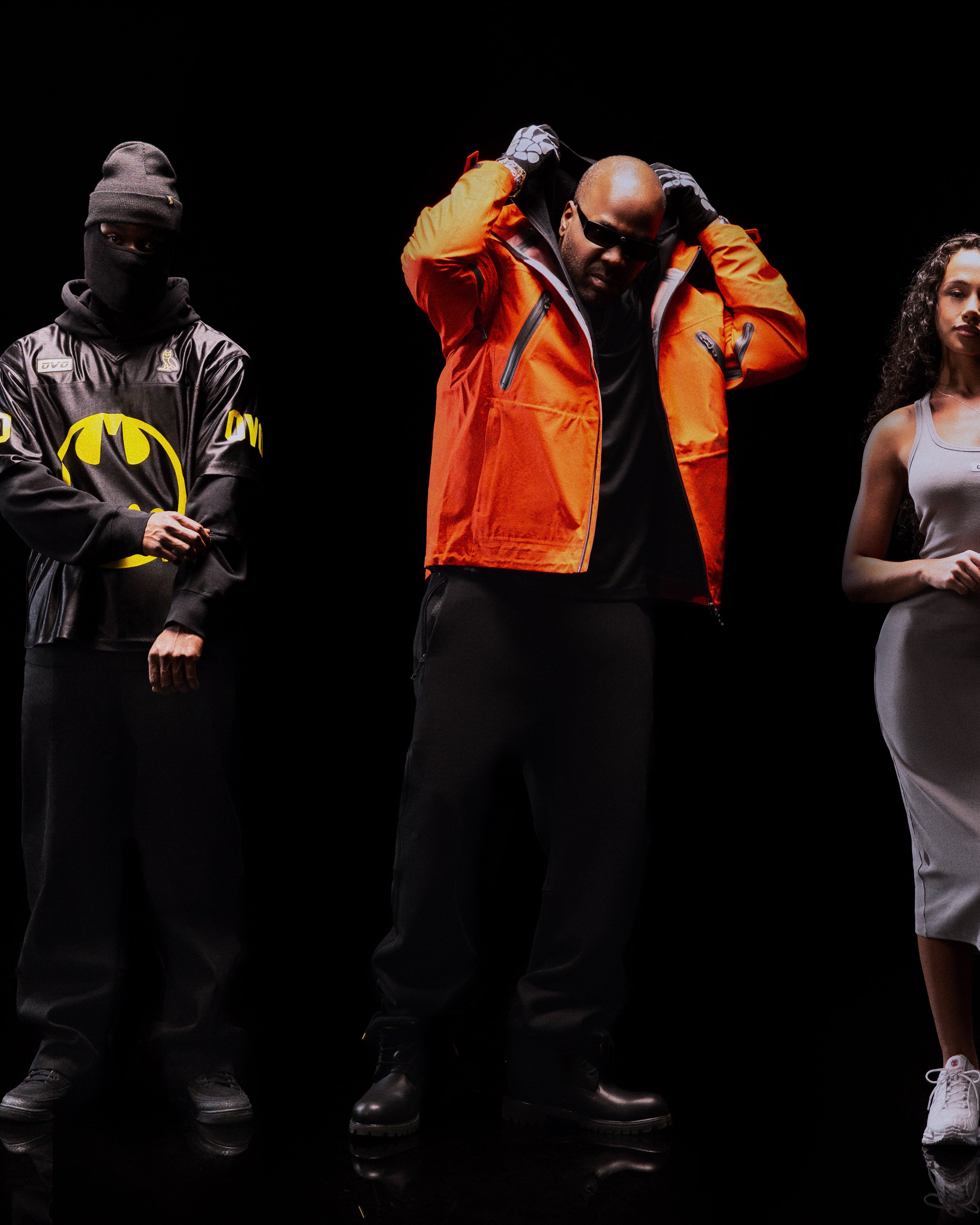 Translation missing: fr.Three people standing against a black background, one in an orange jacket, another in a Batman hoodie, and the third in a white dress
