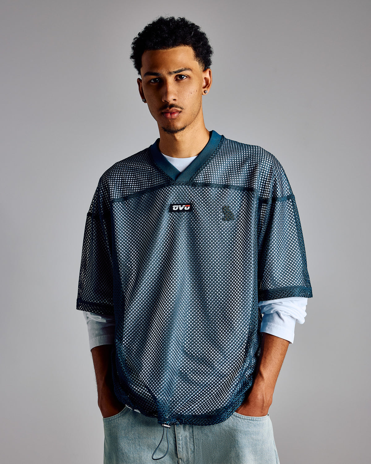 Teal mesh football jersey with dark navy panels, small logo at chest, worn over white long-sleeve shirt.