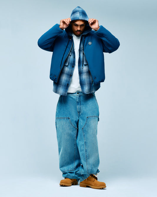 Man wearing blue Primaloft jacket with quilted hood, plaid shirt, white tee, jeans, and tan boots against light blue backgrou