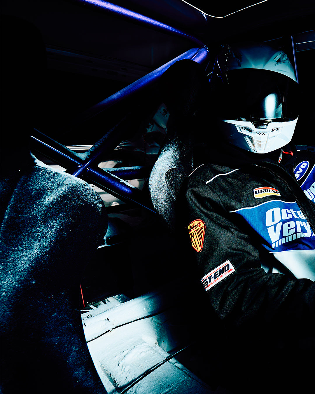 Person in a October's Very Own racing Jacket and helmet inside a car, with visible branding.