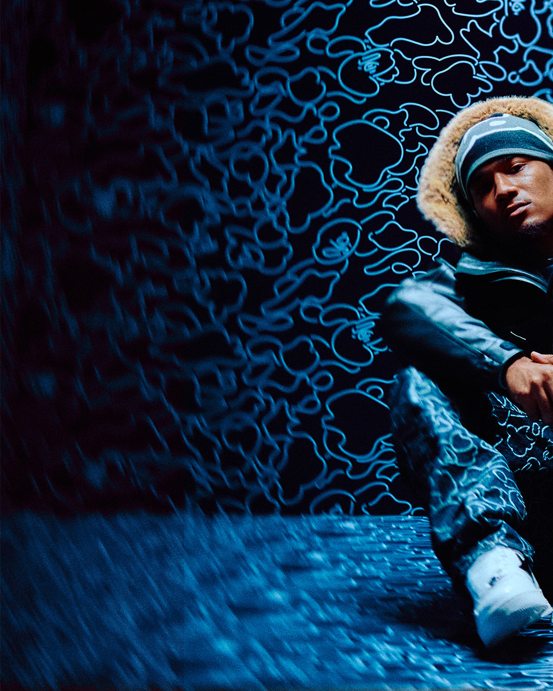 Person wearing October's Very Own x Bape fur-lined coat and hat against a dark, textured wall with abstract patterns.