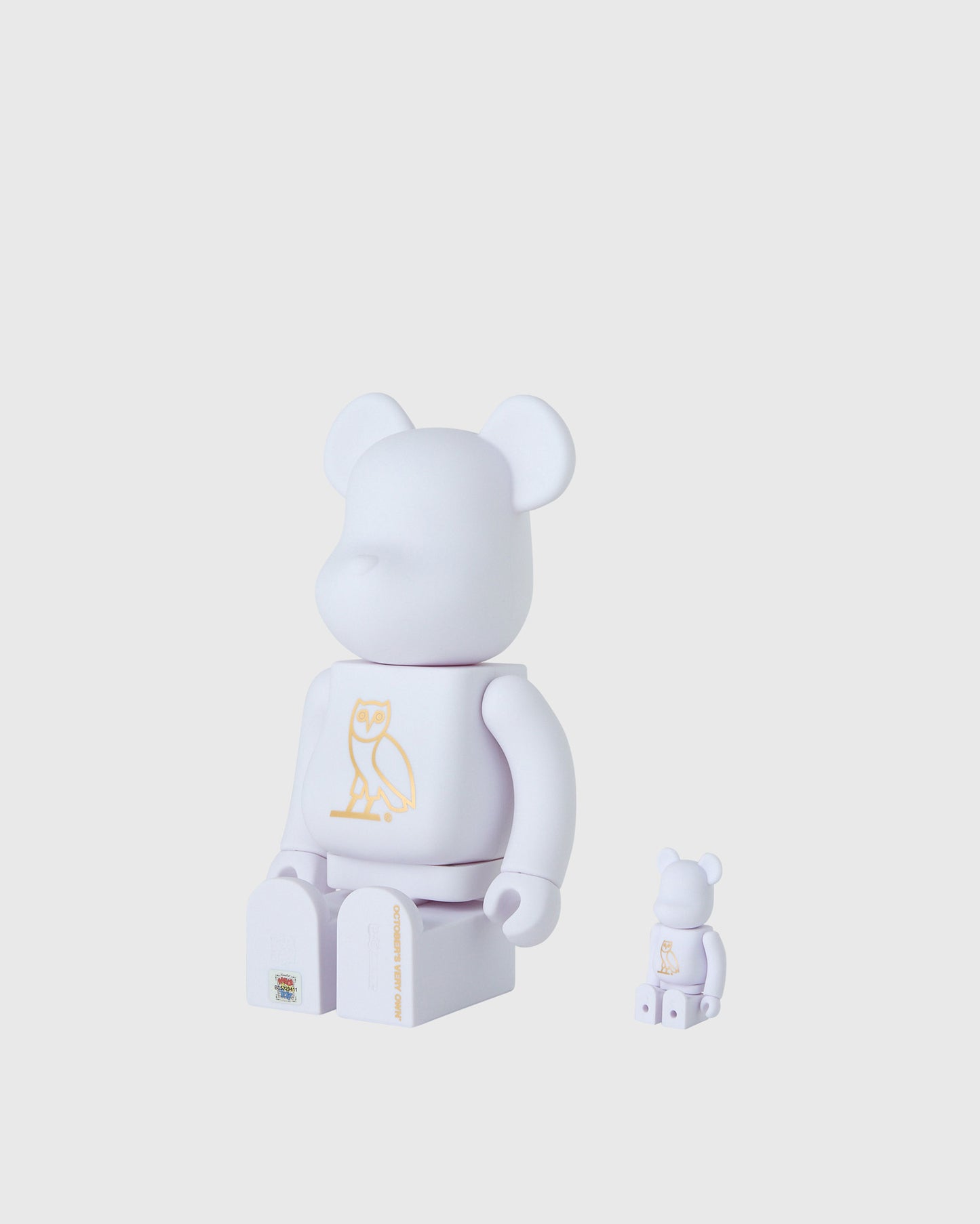 Two pearl white BE@RBRICK figures with rounded ears and gold owl graphic on chest, one larger and one smaller size.