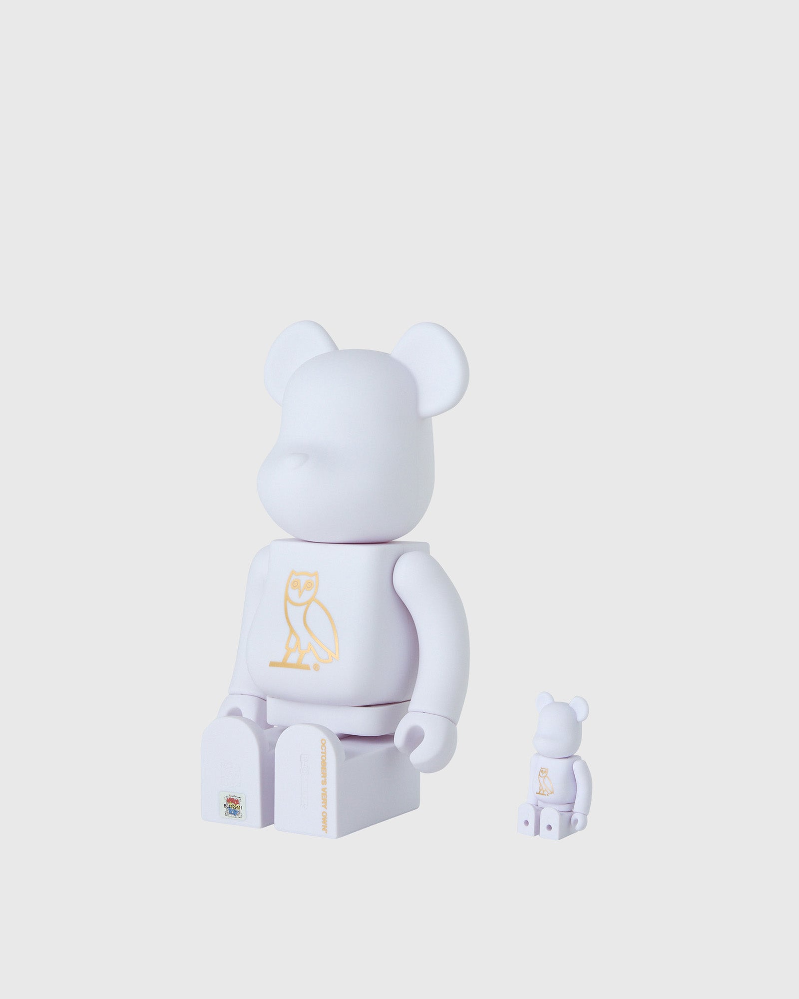 Two pearl white BE@RBRICK figures with rounded ears and gold owl graphic on chest, one larger and one smaller size.