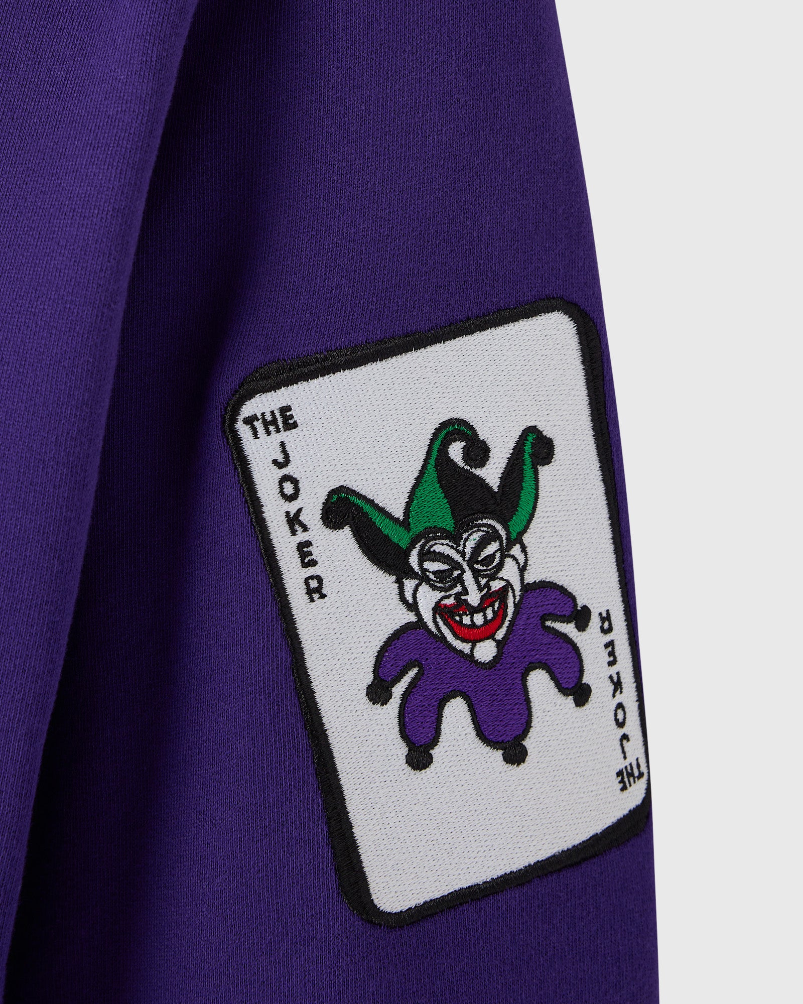 Violet hoodie with embroidered playing card patch featuring Joker character with green hat and purple outfit on chest.