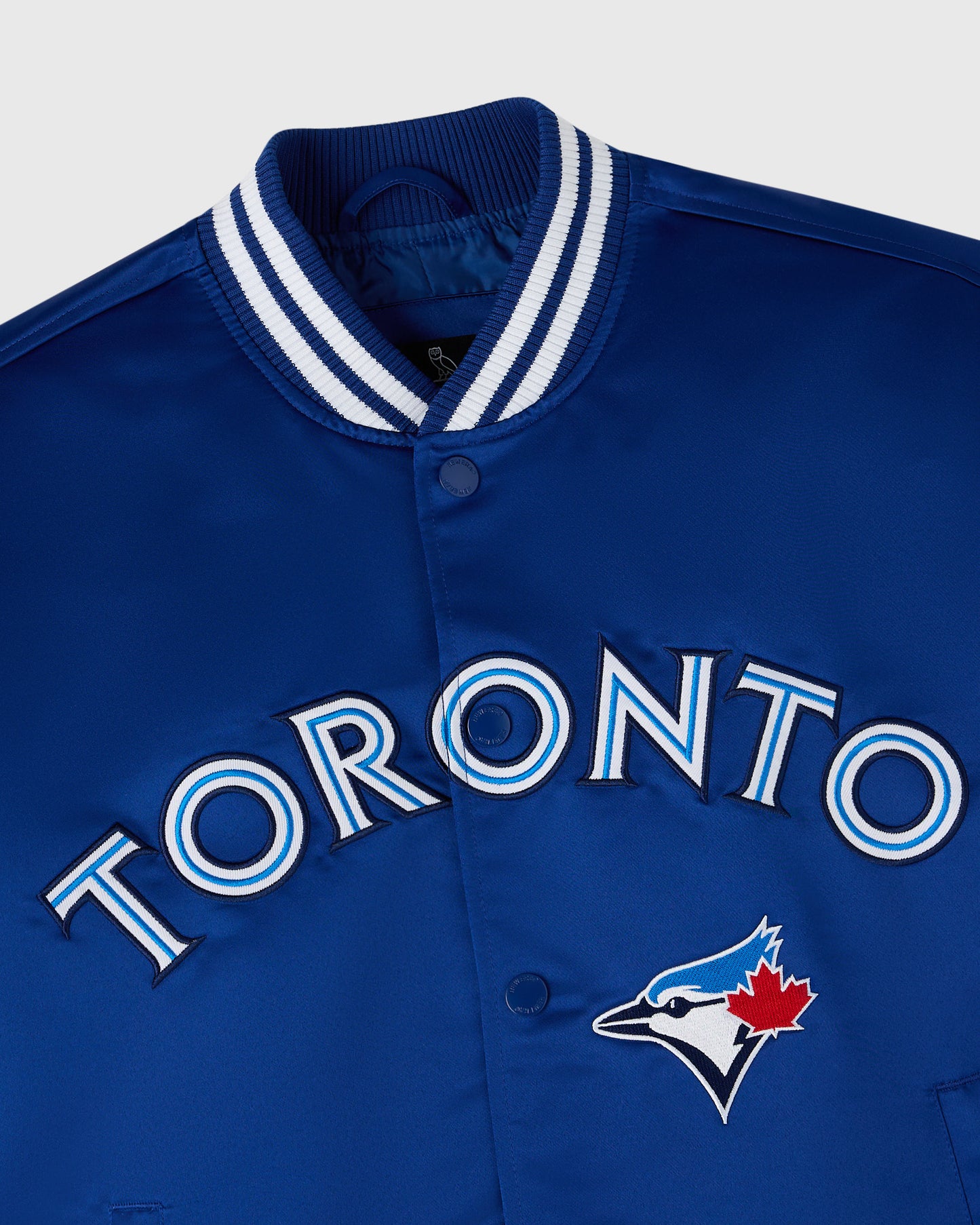 Blue varsity jacket with white and navy striped ribbed collar, "Toronto" text, and Blue Jays bird logo with red maple leaf.