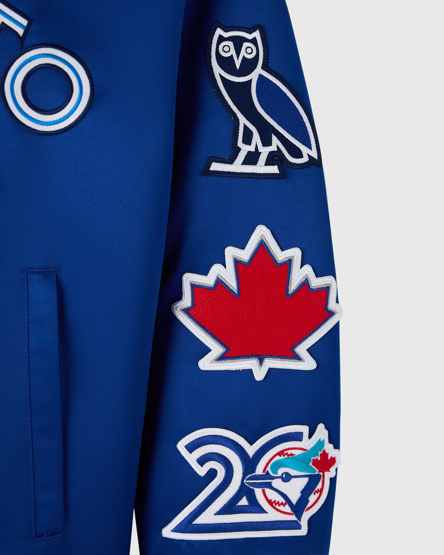 Blue varsity jacket with embroidered owl, maple leaf, and Toronto Blue Jays logos on sleeve.