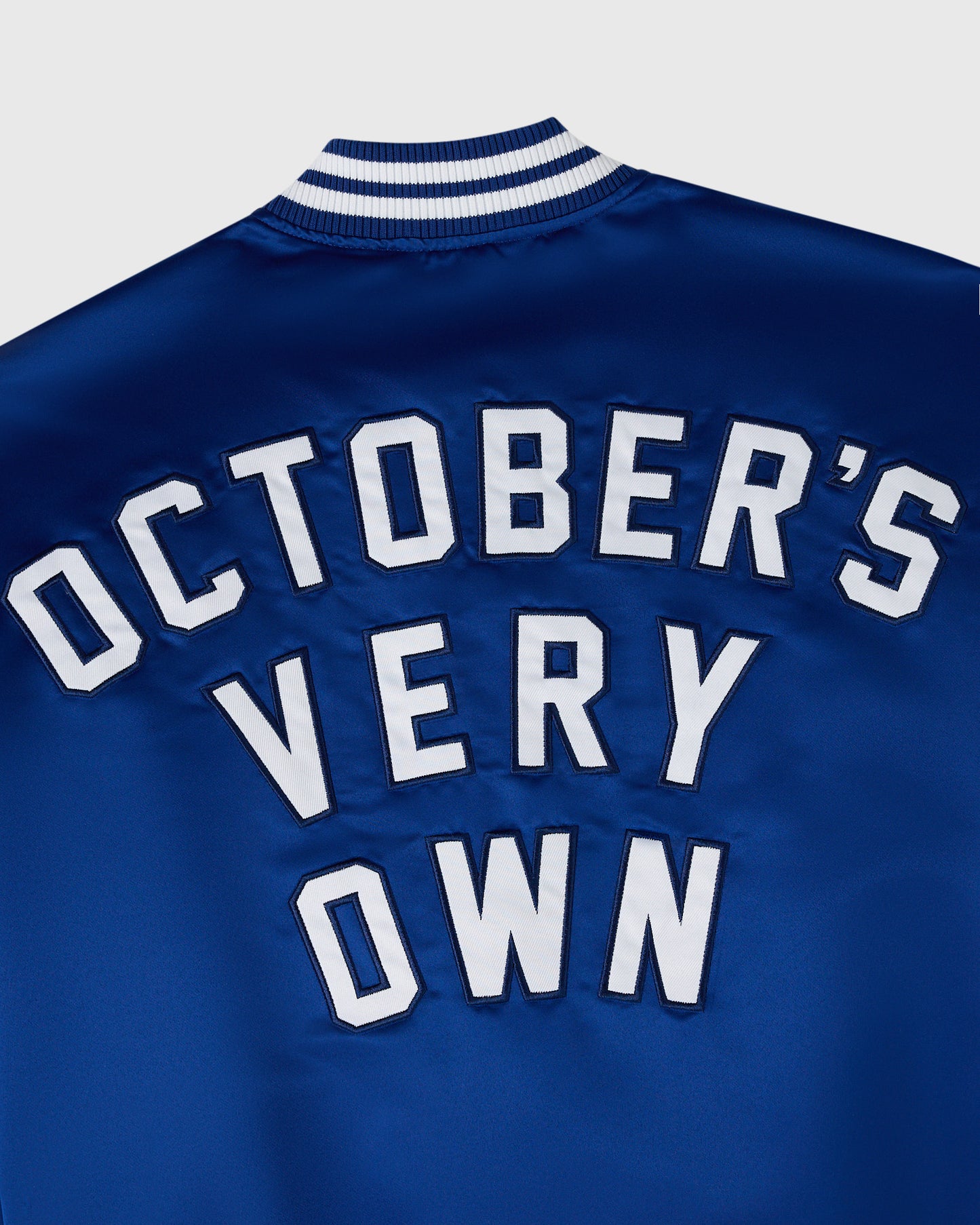 Blue varsity jacket with white "October's Very Own" text on back and navy-white striped ribbed collar.