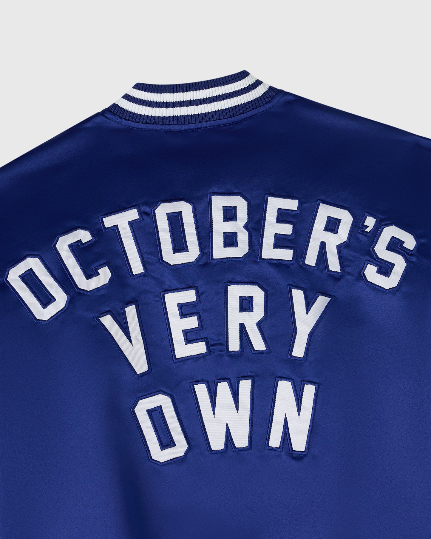 Blue varsity jacket with white striped ribbed neckline and "October's Very Own" text appliqué on chest.