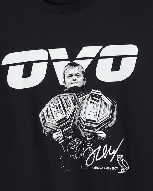 Black t-shirt with white UFC logo and portrait graphic of Hasbulla holding championship belts, signature and name text below.
