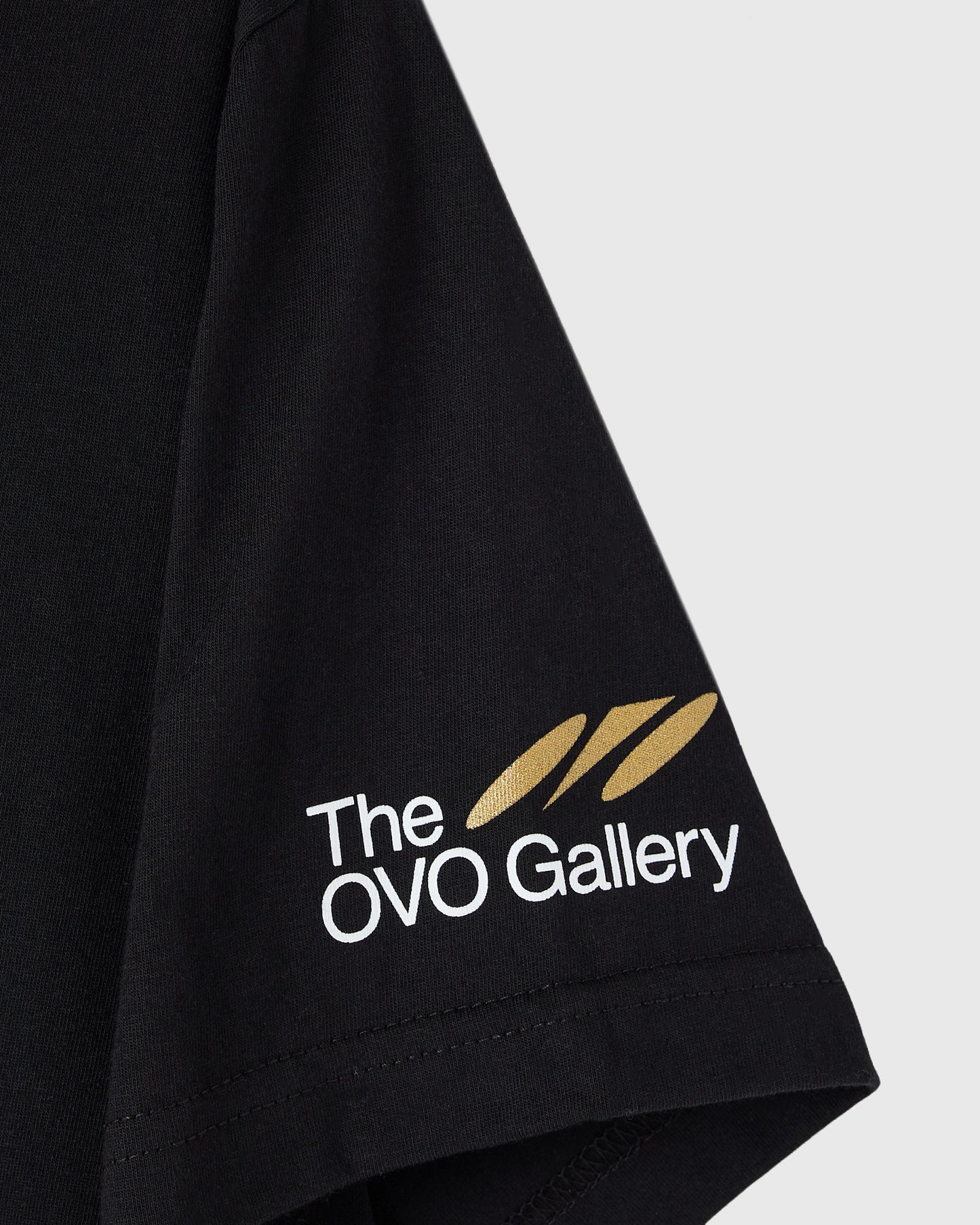 Black t-shirt with "The OVO Gallery" text and gold feather logo printed on sleeve.