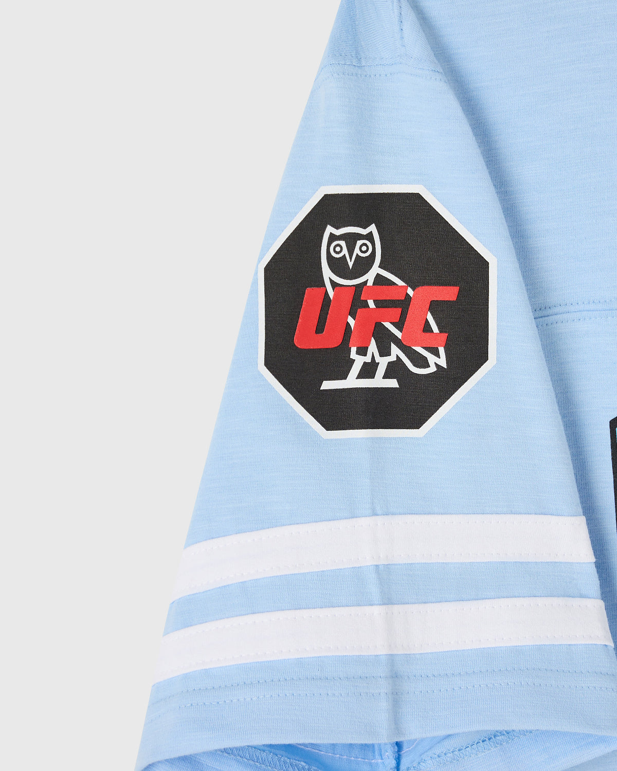 Light blue football tee with white stripes on sleeve and black octagonal UFC Liddell logo patch on chest.
