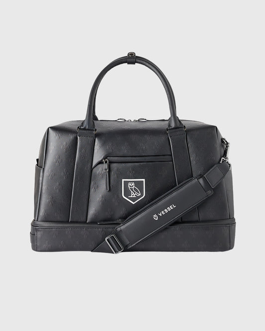 Black Boston bag with dual handles, front zip pocket, monogram shield emblem, and detachable shoulder strap.